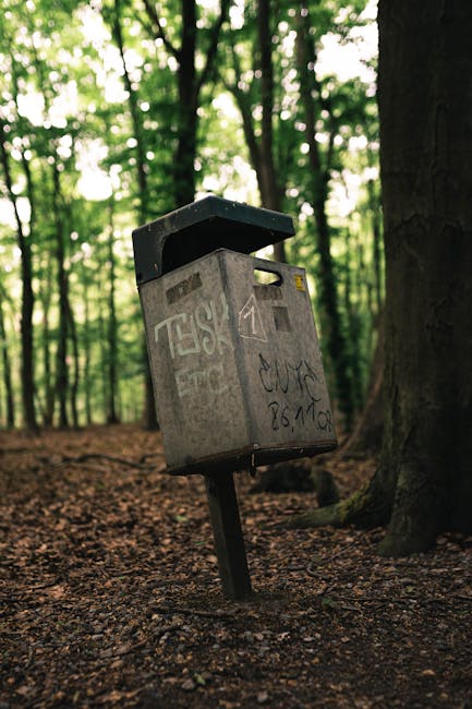 A grey metal bulky waste collection point with graffiti on its surface, mounted on a tilted metal post in a woodland area with tall trees and green foliage in the background. The container is situated