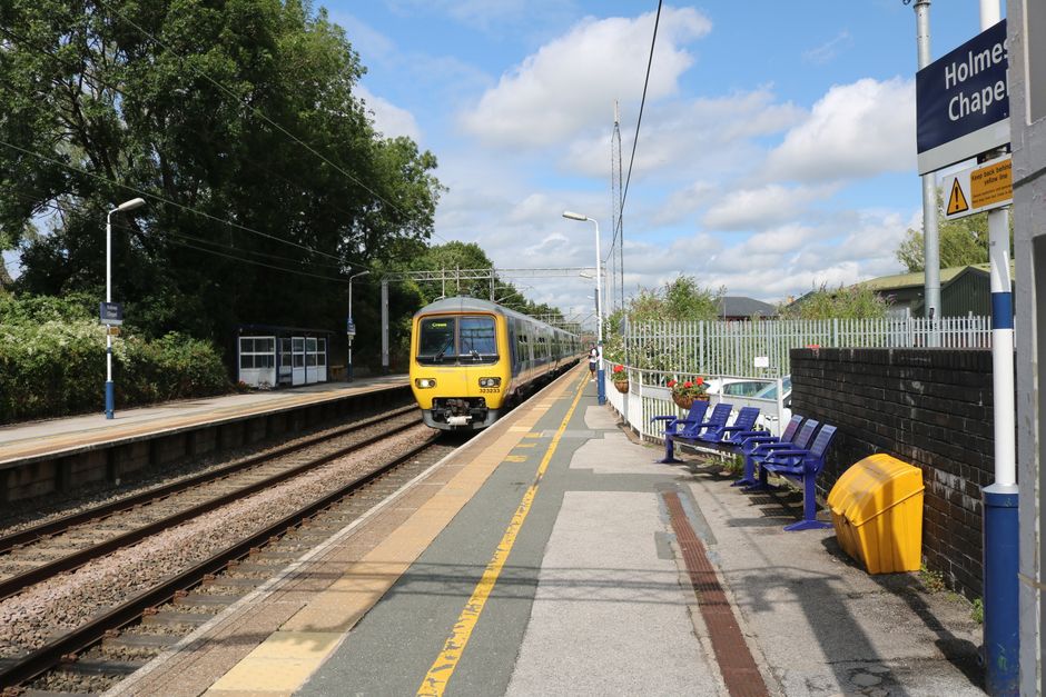 Exterior view of Fulwell Station platform during daytime, featuring a modern white canopy structure supported by curved steel beams casting shadows on the pavement. A train with orange and grey livery