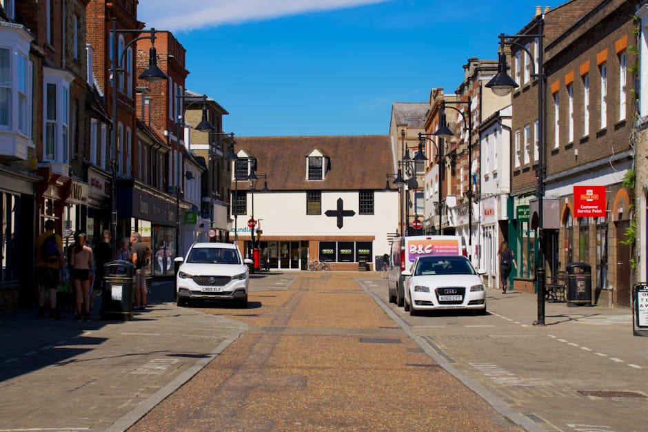 The image shows a pedestrianised street scene in Hampton Hill, with a clear blue sky overhead. On both sides of the street, there are two- to three-storey commercial buildings featuring various shops and retail outlets with large windows and signage. Several people are gathered along the pavement, some standing in small groups, possibly waiting or observing, while others walk or stand near the shop entrances. Parked along the curb are a white van with a ladder on top, a white Audi car, and a small delivery truck with branding visible on the side, indicating local transport services. Street lighting includes black lamp posts with decorative brackets extending over the pavement. In the background, a building with a distinctive cross-shaped window or sign is visible, situated at the end of the street. The scene captures a typical day in an active shopping district, potentially related to home relocation or furniture transport activities as part of a house move or removal project, with [COMPANY_NAME] possibly involved in logistical support for such moves.