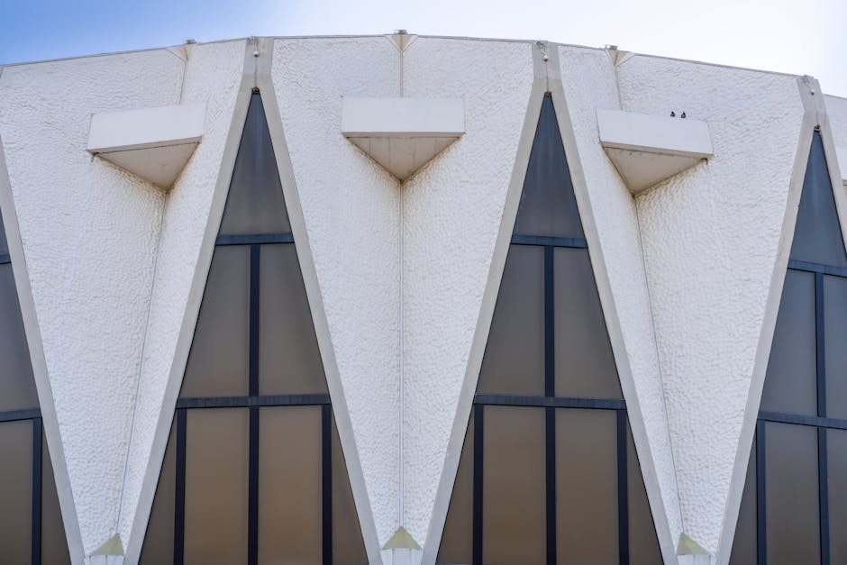 Close-up view of a modern building's exterior featuring three large, V-shaped concrete architectural elements with a textured white surface. Each structure protrudes from the building surface, with small rectangular white concrete balconies at their top. Behind these structures, there are tall, narrow windows divided into multiple panes by black framing, set within a dark facade. The image is taken during daylight with natural lighting highlighting the geometric design. This architectural detail is part of a commercial or institutional property, relevant to house removals and home relocation services offered by Man with Van Hampton Hill, illustrating building features encountered during furniture transport and loading processes for a move.