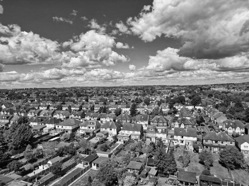 Black and white aerial photograph of a residential neighbourhood showing rows of detached and semi-detached houses with pitched roofs, neatly arranged gardens, and narrow streets interspersed with trees. The image captures the surrounding suburban environment, with some properties having visible fences, lawns, and outdoor storage sheds. In the foreground, rooftops and backyards are clearly visible, indicating a densely populated area. The sky above features scattered fluffy clouds, creating a sense of depth and open space. This scene reflects a typical UK suburban setting suitable for house removals and home relocation services, with Man with Van Hampton Hill potentially operating in such environments, offering professional transport and packing solutions suitable for moving households and furniture within residential areas.