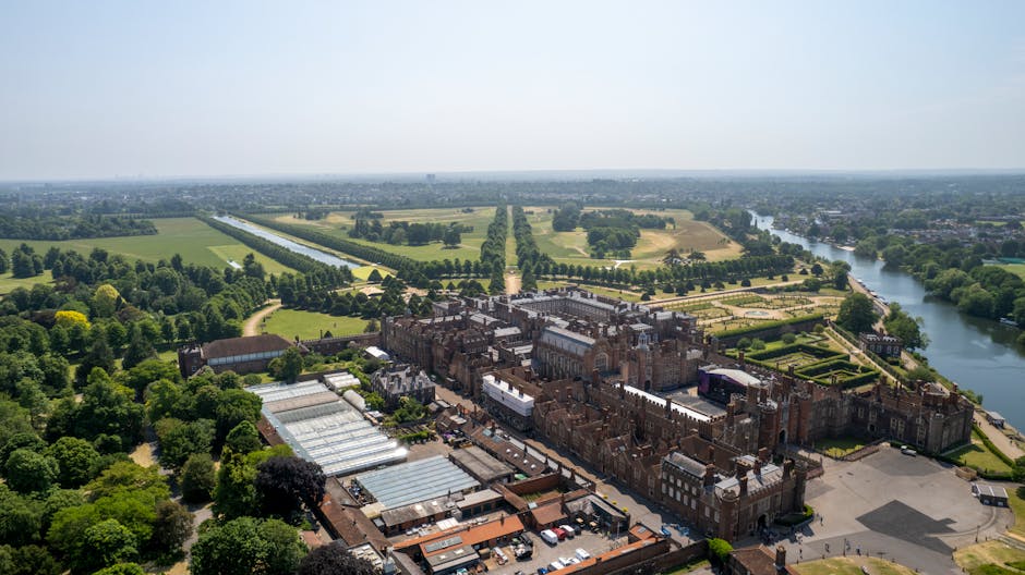 An aerial view of Hampton Court Palace surrounded by extensive green gardens, tree-lined courtyards, and adjacent parking areas, with a river on the right side. In the foreground, there are several glass-roofed structures within the palace grounds. The scene is captured during daylight with clear weather. This image reflects the historic site often involved in house removals and moving logistics for residents and visitors. Man with Van Hampton Hill provides house removals and packing services, supporting relocation projects in the area and ensuring smooth furniture transport for clients. The palace's exterior, lush landscaping, and accessible transport routes highlight the environment for home relocation and moving services in Hampton Hill, with the surrounding grounds serving as a staging area for loading and unloading furniture and packed boxes during property moves.