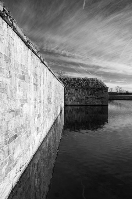 A black-and-white photograph captures a scene near a waterway, featuring a large stone wall running along the edge of the water. The wall is constructed from neatly arranged rectangular stones and ext