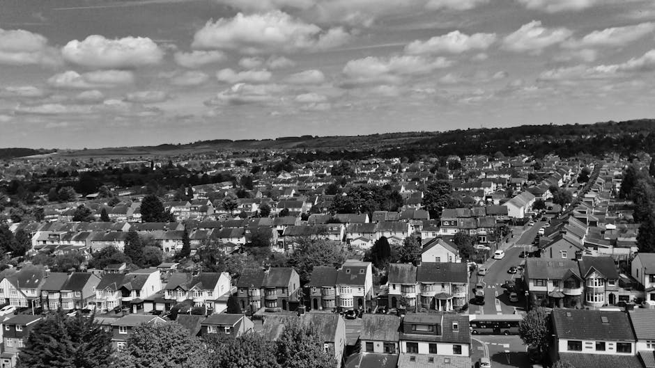 A black-and-white aerial view of a residential suburb showing rows of closely packed terraced houses and detached homes along streets, with trees interspersed between the properties. The scene captures a wide expanse of the neighborhood, with some vehicles parked along the roads. The sky above is partly cloudy. This image, associated with house removals and furniture transport, illustrates the typical environment where local moving services like those from Man with Van Hampton Hill operate, highlighting the suburban streets and housing layout involved in home relocation processes.