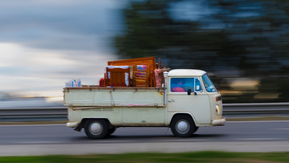 A vintage white van with a rounded front and side mirror, positioned on a blurred road to indicate motion, is loaded with various household furniture and packing items. Inside the van's open flatbed, there are wooden chairs, cardboard boxes, and plastic containers, all secured with packing materials. The furniture appears to be a mix of dining chairs and small tables, wrapped in fabric or blankets for protection during transport. The scene is set outdoors on a road with a highway barrier visible behind, and the background features a blurred landscape of trees and sky, suggesting the vehicle is in transit. The image reflects the process of home relocation, with furniture and belongings staged for moving, aligning with services offered by Man with Van Hampton Hill for furniture transport and packing during house removals.