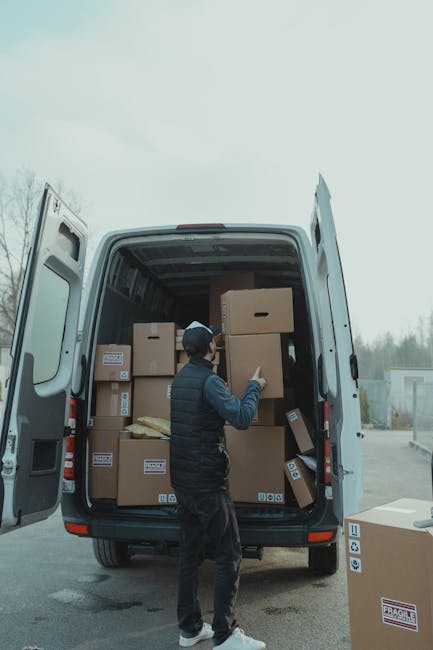 A man wearing a dark jacket, black trousers, and a black cap is standing outside next to the open rear doors of a large white van, which is parked on a paved surface. Inside the van, numerous cardboard boxes of different sizes are visible, some secured with plastic wrap or packing tape, and a few are stacked neatly while others are slightly tilted. The man appears to be in the process of loading or organizing the boxes, with his right hand holding a smaller box positioned near the larger boxes in front of him. The back doors of the van are wide open, revealing the interior filled with additional packed boxes and some packaging materials. The setting suggests a home relocation or moving process, supported by the presence of a single cardboard box on the ground beside the vehicle, marked as fragile. The background features a cloudy sky and a faint view of trees and structures in the distance, indicating an outdoor environment, possibly near a residential or commercial property. This scene exemplifies the loading process involved in professional removals services, such as those provided by Man with Van Hampton Hill, facilitating efficient furniture transport and packing during house moves in Hampton Hill.
