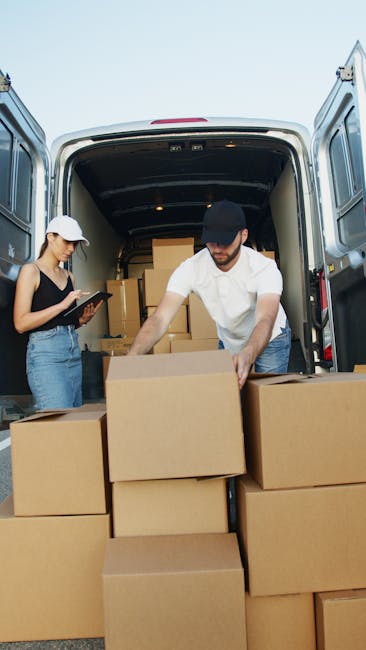 Two individuals are engaged in a home relocation process involving a van parked outdoors. The man, wearing a white shirt and a black cap, is actively loading large cardboard boxes into the back of the vehicle, which is open and filled with more boxes and packaging materials. The woman, dressed in a black tank top, light blue jeans, and a white cap, is standing nearby holding a tablet, possibly coordinating the packing or coordination details. The boxes are stacked inside the van and outside on the pavement, some still on the ground awaiting transfer. The van's interior shows a spacious cargo area with visible cardboard boxes and packing materials, indicating a professional furniture transport and moving process. Natural daylight illuminates the scene, supporting a clear view of the loading activity. This image illustrates the logistics involved in house removals and packing services provided by companies like Man with Van Hampton Hill, supporting professional small business moves in Hampton Hill.