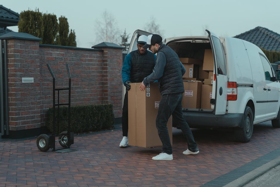 A man wearing a dark jacket, black trousers, and a black cap is standing outside next to the open rear doors of a large white van, which is parked on a paved surface. Inside the van, numerous cardboard boxes of different sizes are visible, some secured with plastic wrap or packing tape, and a few are stacked neatly while others are slightly tilted. The man appears to be in the process of loading or organizing the boxes, with his right hand holding a smaller box positioned near the larger boxes in front of him. The back doors of the van are wide open, revealing the interior filled with additional packed boxes and some packaging materials. The setting suggests a home relocation or moving process, supported by the presence of a single cardboard box on the ground beside the vehicle, marked as fragile. The background features a cloudy sky and a faint view of trees and structures in the distance, indicating an outdoor environment, possibly near a residential or commercial property. This scene exemplifies the loading process involved in professional removals services, such as those provided by Man with Van Hampton Hill, facilitating efficient furniture transport and packing during house moves in Hampton Hill.