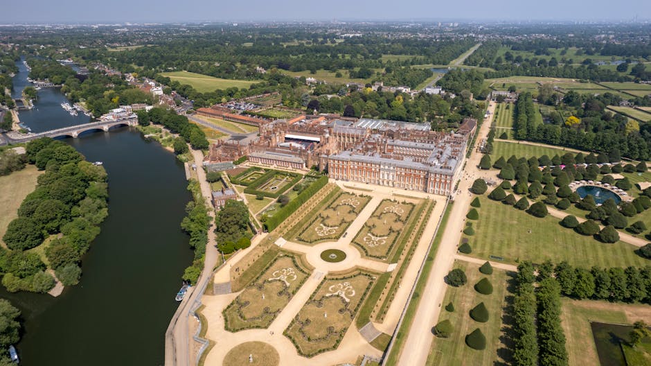 An aerial view of a large historic mansion with ornate gardens, landscaped lawns, and symmetrical flower beds, situated near a river with a bridge crossing over it. The mansion has a grand, classical architecture style with multiple chimneys, and the surrounding grounds feature neatly trimmed trees and pathways. In the foreground, a team from Man with Van Hampton Hill is seen loading furniture wrapped in protective blankets and plastic wrap onto a van parked on a paved driveway adjacent to the property, indicating a home relocation or furniture transport process. The vibrant greenery and open space reflect the spacious setting typical of high-end house removals and packing and moving services near Bushy Park, Hampton Hill.