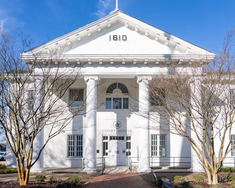 The front view of a white, neoclassical-style court house building with the street number 1610 displayed on the triangular pediment above the entrance. The structure features large white columns supporting the decorative cornice, with a central arched window above the double doors that have small glass panels. The building is framed by leafless trees on either side, with bare branches extending across the façade, and sunlight casting shadows on the white exterior. The surrounding area includes a paved walkway leading up to the entrance, and a low black metal railing along the steps. The clear blue sky in the background indicates a bright, sunny day. This image could be relevant for illustrating property or building exteriors in the context of house removals or physical relocation services provided by companies like Man with Van Hampton Hill.