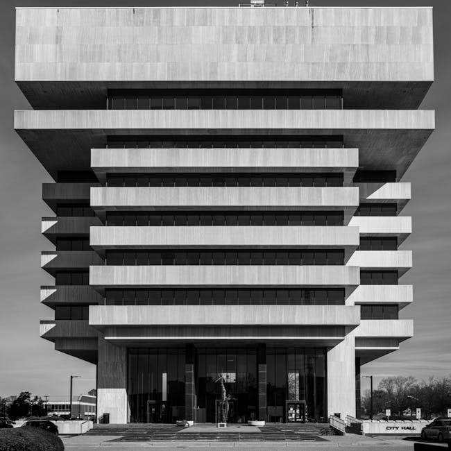 A black and white photograph of a modern multi-storey building, identified as city hall, featuring a distinctive architectural design with large, protruding horizontal concrete slabs creating a layered appearance. The building's facade has extensive glass windows at the ground level, providing a view into the interior lobby area, and the entrance is accessed via a short set of steps. Surrounding the building are paved pathways, some street furniture, and a few trees in the background under a partly cloudy sky. The image captures the structure's imposing presence and geometric detailing, with clear daytime lighting highlighting the sharp lines and textures of the concrete surfaces. This type of architecture is relevant to property and building management services, including the logistical aspects of home relocation or commercial moving projects, which companies like Man with Van Hampton Hill may assist with when planning furniture transport and packing during a house removal or office move.